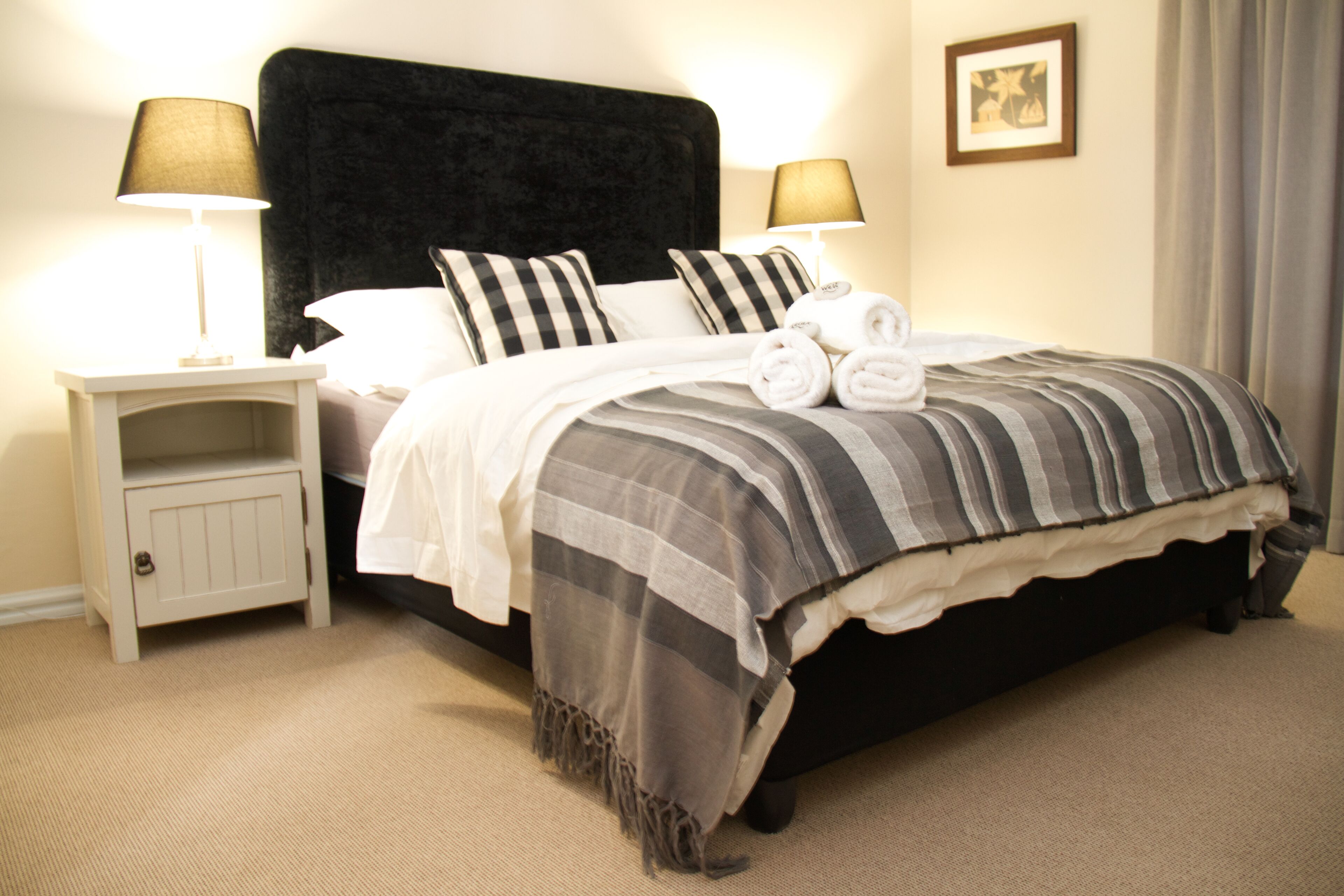 A neatly made bed with a striped blanket and two pillows, two bedside lamps, a small cabinet, and a framed picture on the wall.