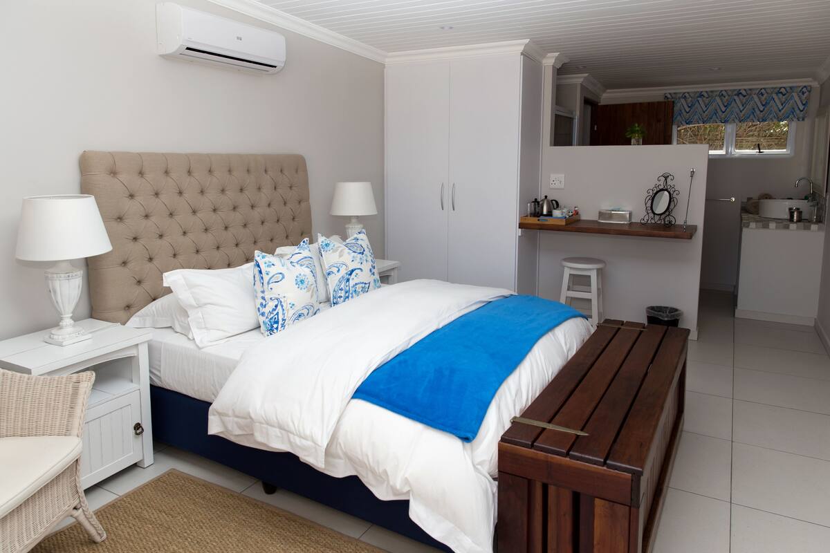 A bedroom with a bed, bedside tables, a lamp, a bench, and a view into a kitchen area.