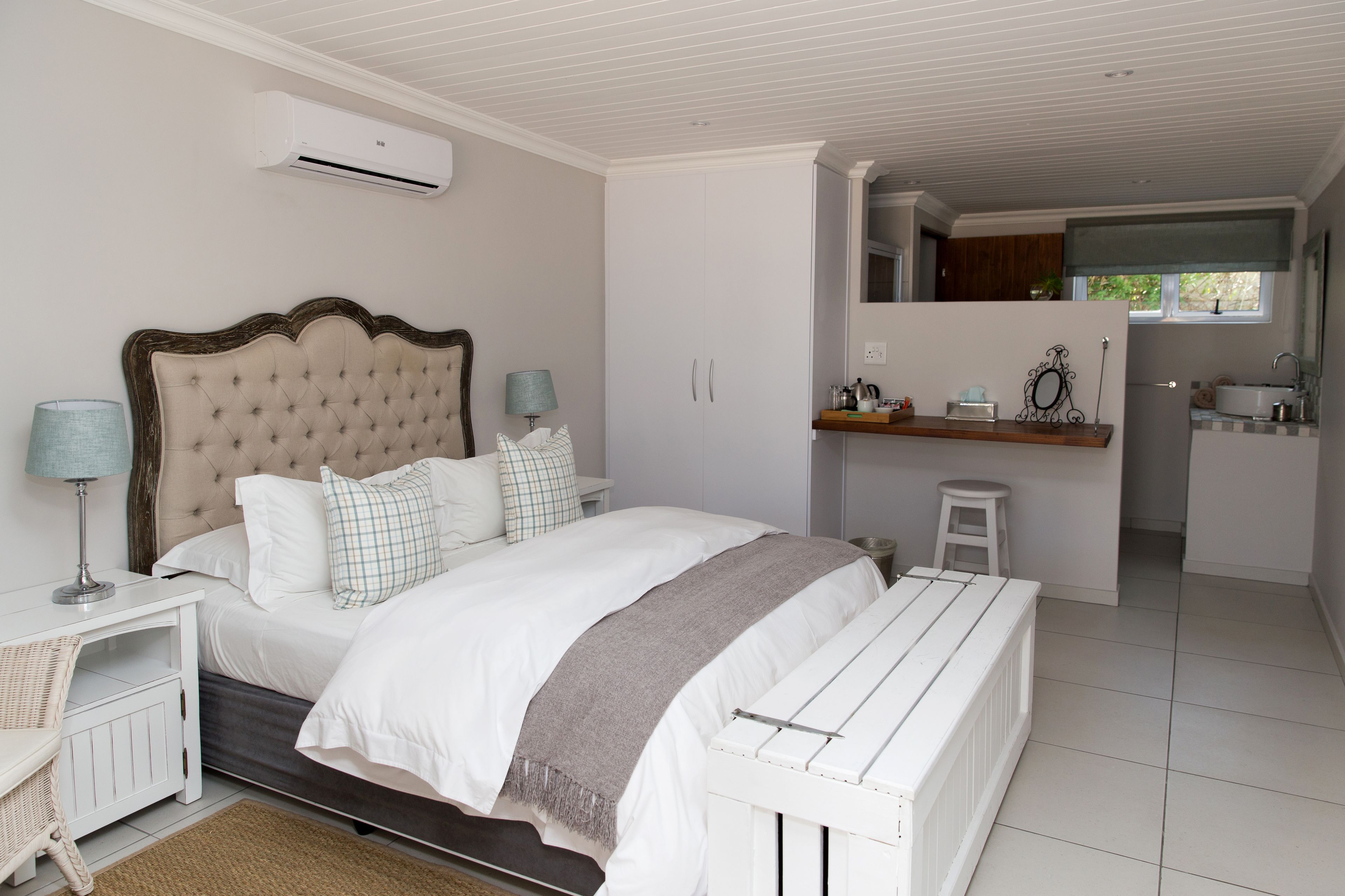 Un dormitorio con una cama grande, mesitas de noche, un taburete y una silla. Incluye una kitchenette con un lavamanos y una ventana.