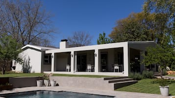 Relax in a spacious patio area surrounded by a well-maintained lawn and mature trees, with a large swimming pool and comfortable seating.
