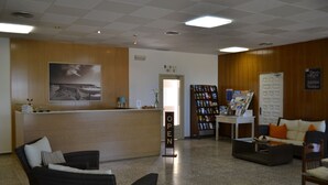 A reception area with a wooden reception desk, a waiting area with chairs, a bookshelf, and a sign indicating the rooms are available. - Hostal L´Esplai (Viladamat)