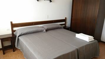 Una cama bien hecha con colcha y almohadas grises, una mesita de noche de madera, un armario de madera y un gancho fijado a la pared.
