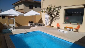 A swimming pool with lounge chairs and umbrellas, surrounded by a building with a tiled roof and a tree with yellow flowers. - Hostal L´Esplai (Viladamat)