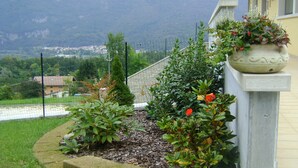 A landscaped garden with a stone border, a potted plant on a pedestal, and a view of a town and mountains in the background. - Affittacamere Rubino (Ponte nelle Alpi)