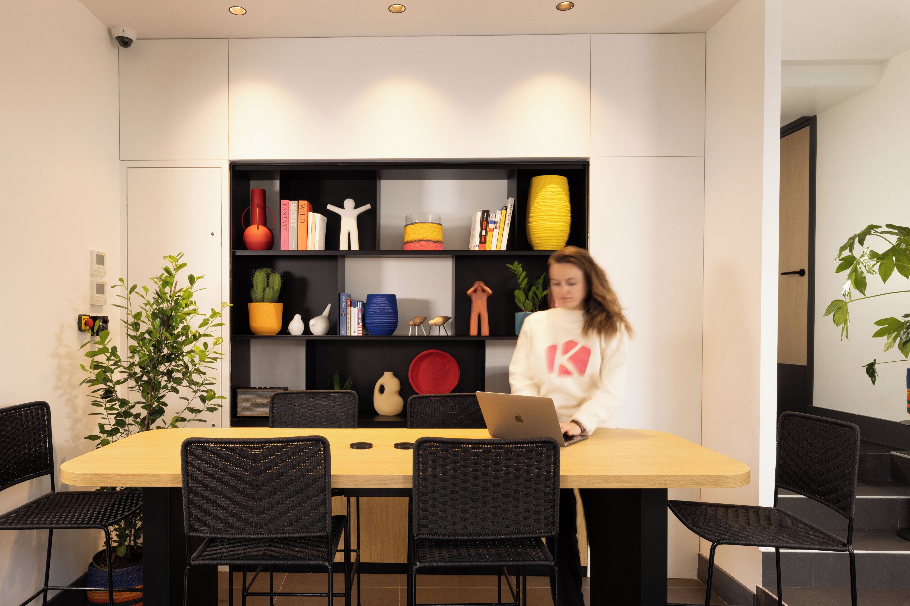 Un comedor moderno con una mesa de madera y sillas negras, una estantería con diversos objetos decorativos, y una planta en maceta.