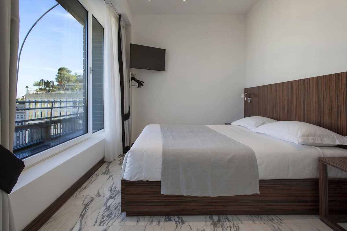 A hotel room with a large window, a bed with white linens, a wooden headboard, a TV mounted on the wall, and a marble floor.