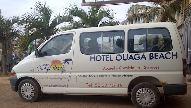 Un fourgon blanc avec l’inscription « HÔTEL OUAGA BEACH » et les coordonnées de l’établissement sur le côté.