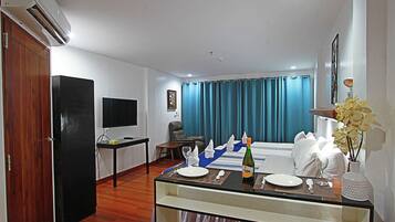 Habitación de hotel con comedor, televisor, escritorio, silla, cama, ventana con cortinas y un jarrón con flores.