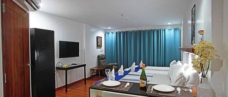 Habitación de hotel con comedor, televisor, escritorio, silla, cama, ventana con cortinas y un jarrón con flores.