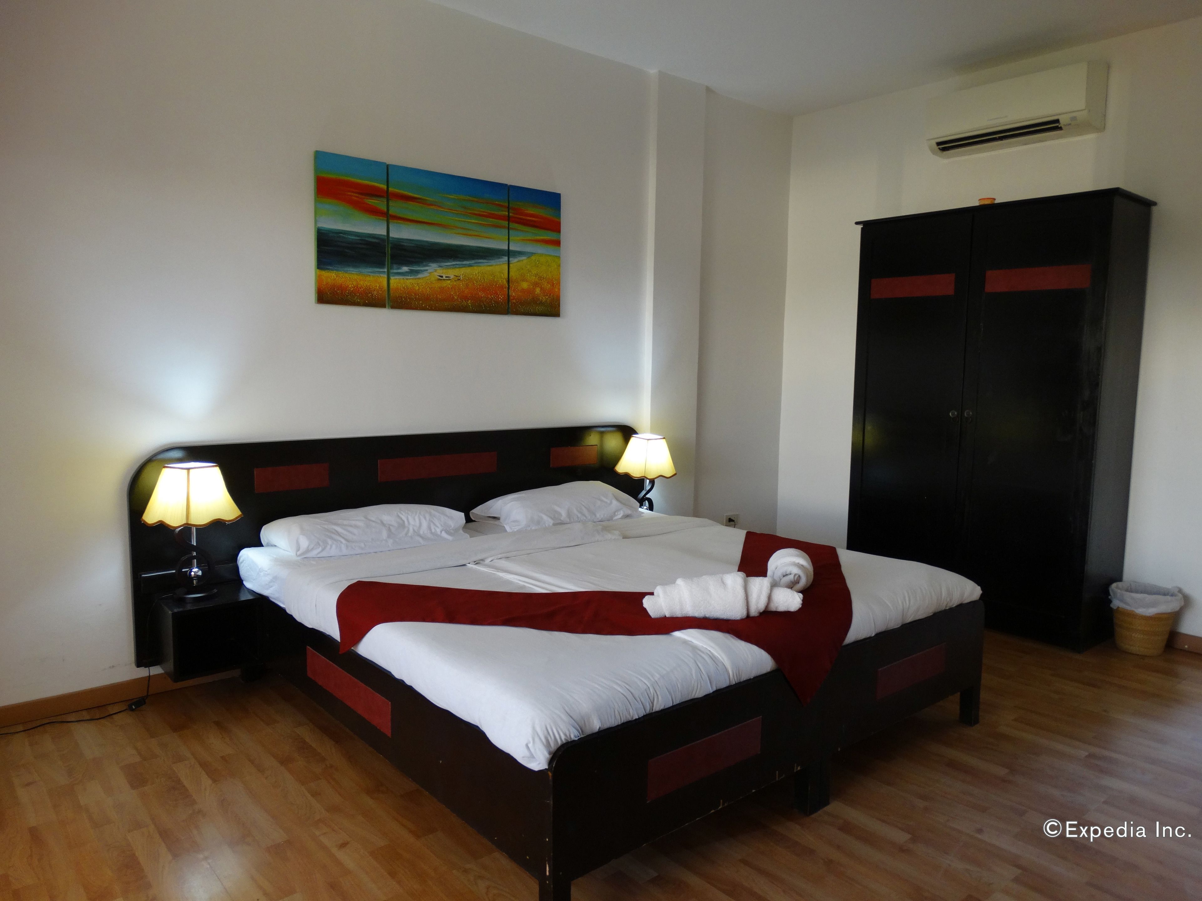 A hotel room with a bed, two bedside lamps, a painting on the wall, a wardrobe, and a trash bin.