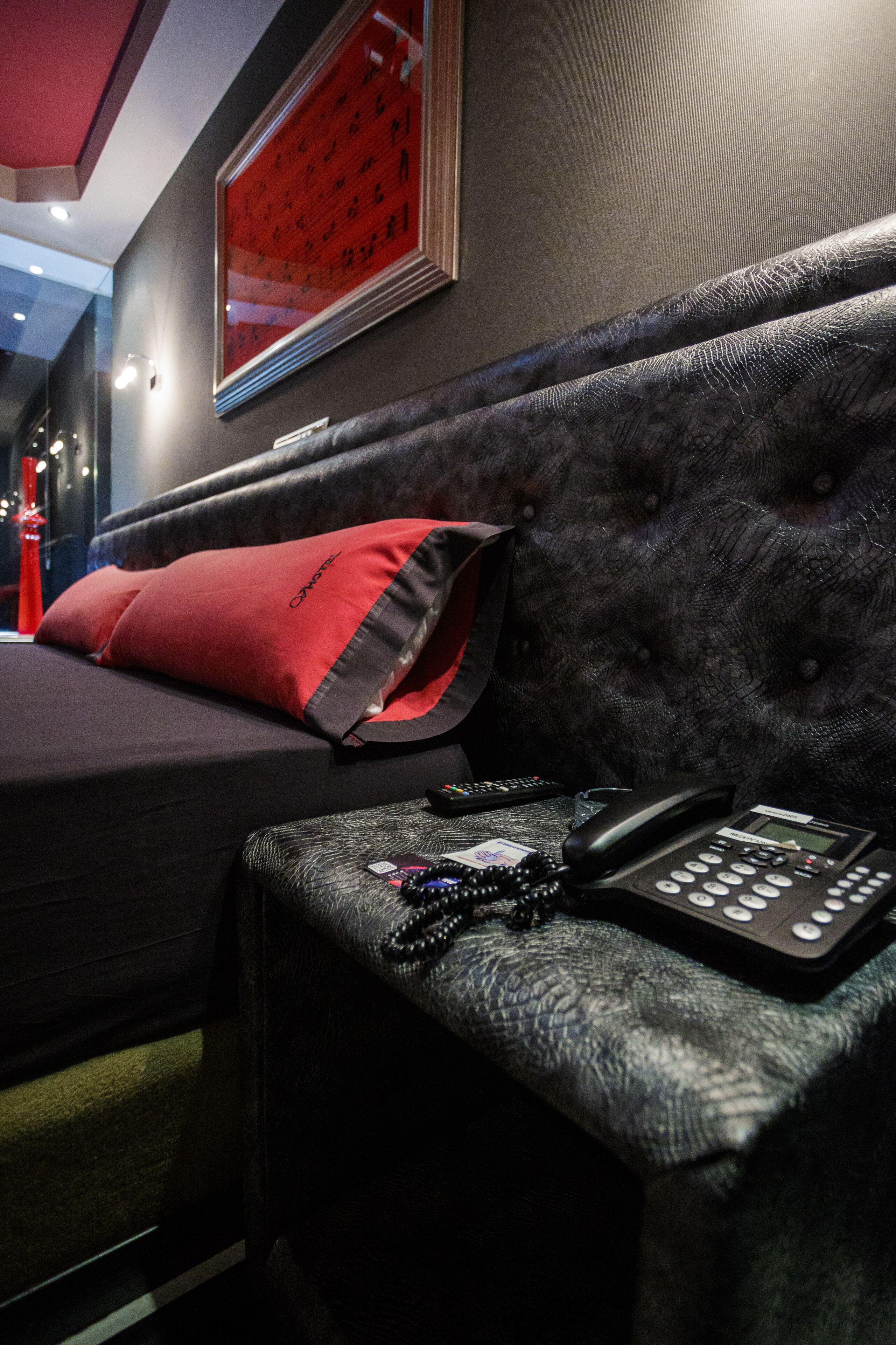 A hotel room with a dark upholstered headboard, a red pillow, a black telephone, and a framed artwork on the wall.