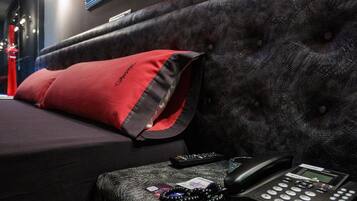 A hotel room with a dark upholstered headboard, a red pillow, a black telephone, and a framed artwork on the wall.