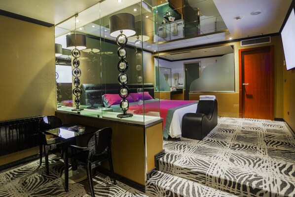 A hotel room with a glass partition, a bed with a purple blanket, a desk with a computer, a chair, and a zebra-patterned carpet. - Motel Venus Pontevedra (Poio)