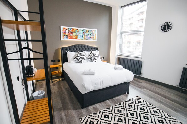 A modern bedroom with a large bed, a black headboard, a colorful artwork above the bed, a black clock on the wall, and a window with blinds. - The Black Lion Manchester (Salford)