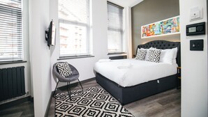 A modern bedroom with a large bed, a black and white patterned rug, a grey chair, and a colorful artwork on the wall. - The Black Lion Manchester (Salford)
