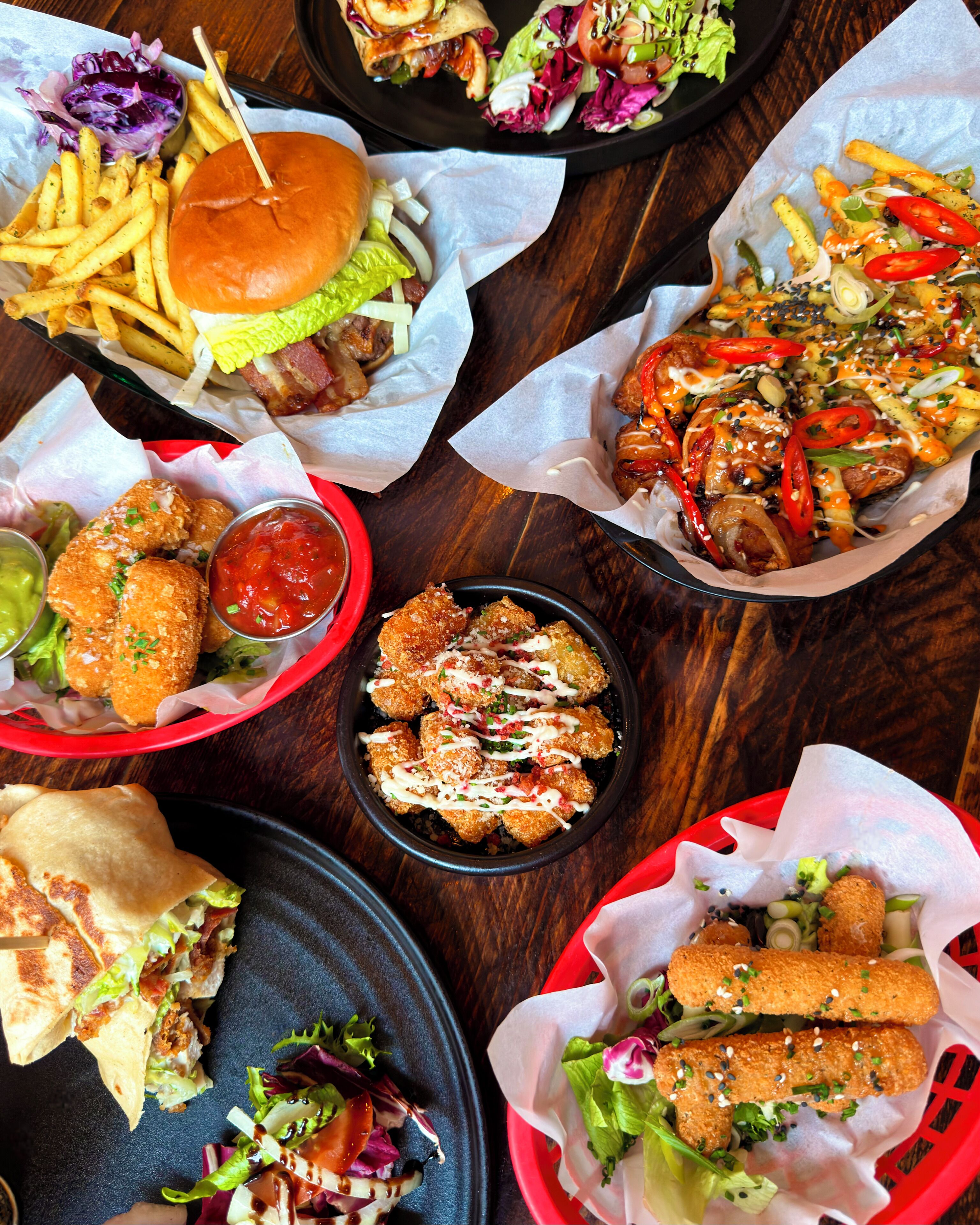 Variedad de alimentos, incluyendo hamburguesas, papas fritas y alitas de pollo, sobre una mesa de madera.