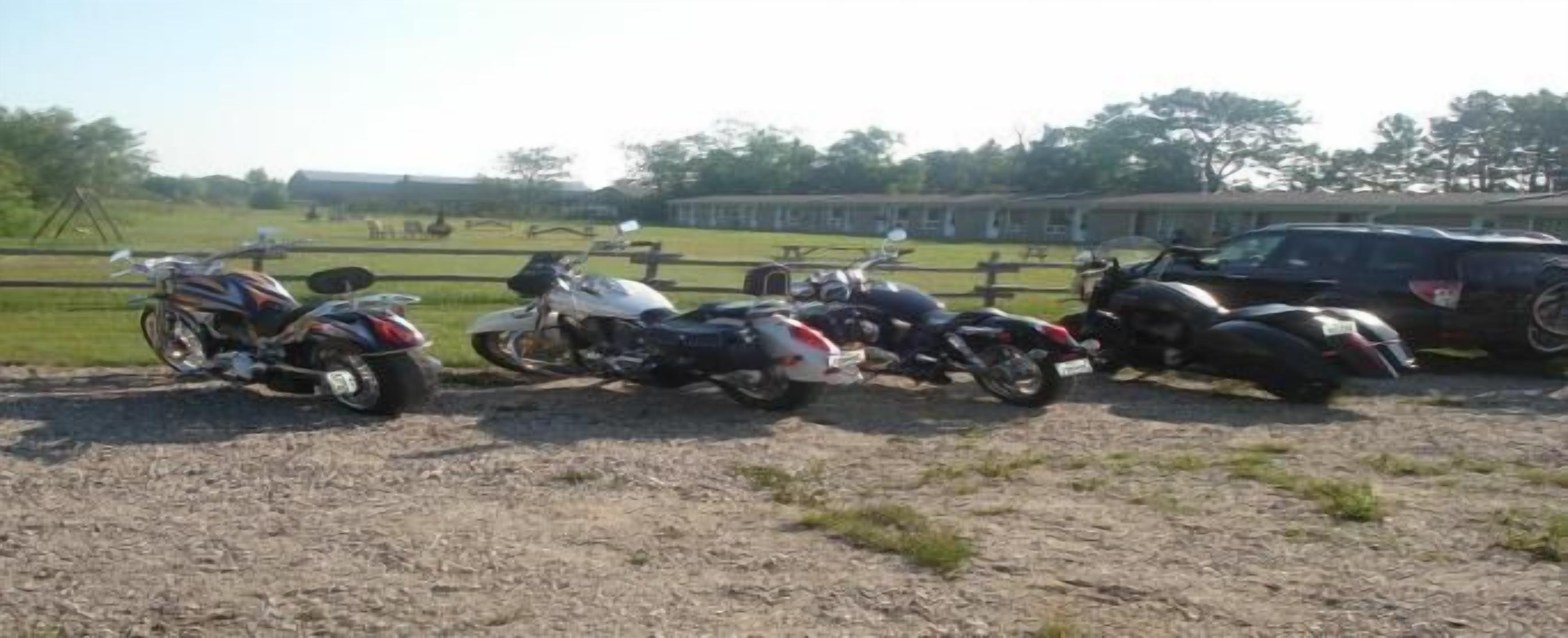 Tres motocicletas estacionadas en un área de grava, con una valla y árboles al fondo.