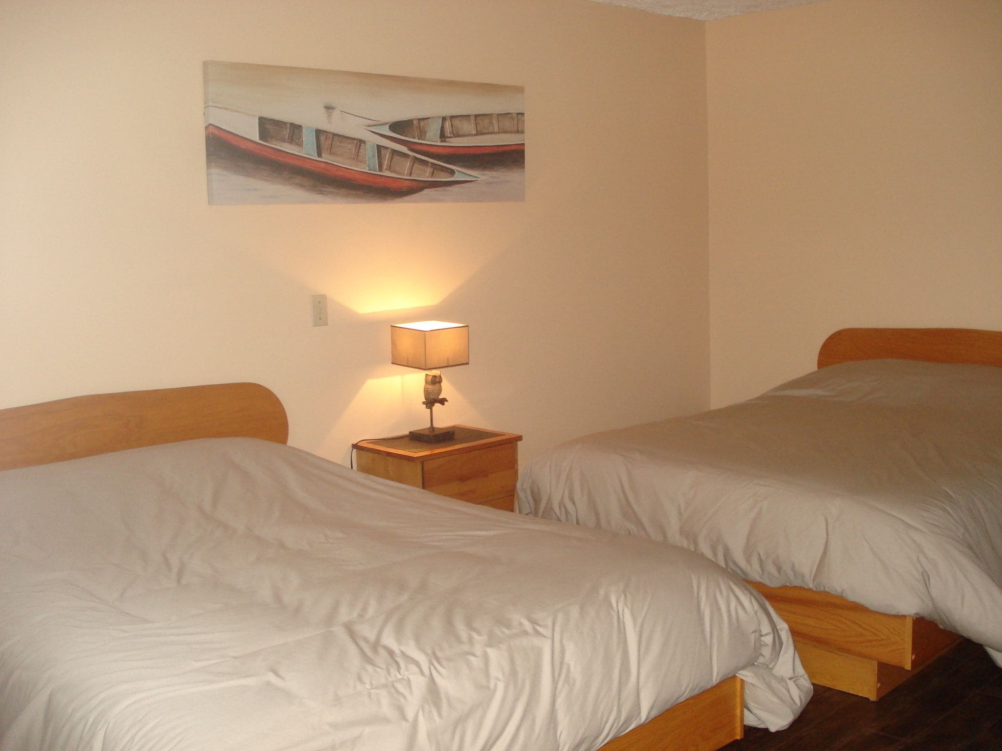 A hotel room with two beds, a nightstand with a lamp, and a painting of boats on the wall.