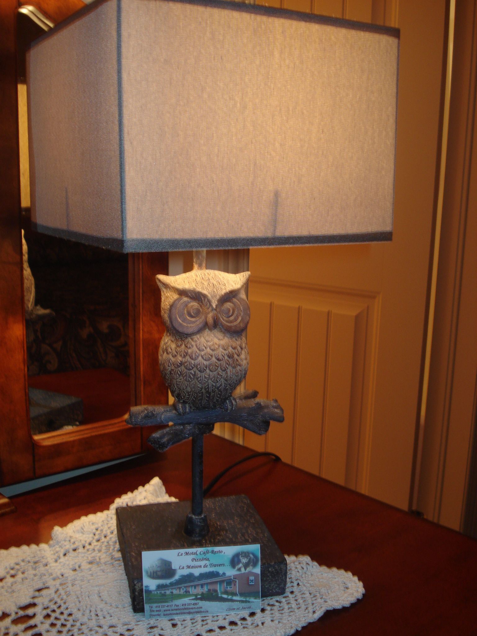 Une lampe dont le pied est en forme de hibou et l’abat-jour rectangulaire, posée sur une table en bois avec une petite plaque fixée en dessous.