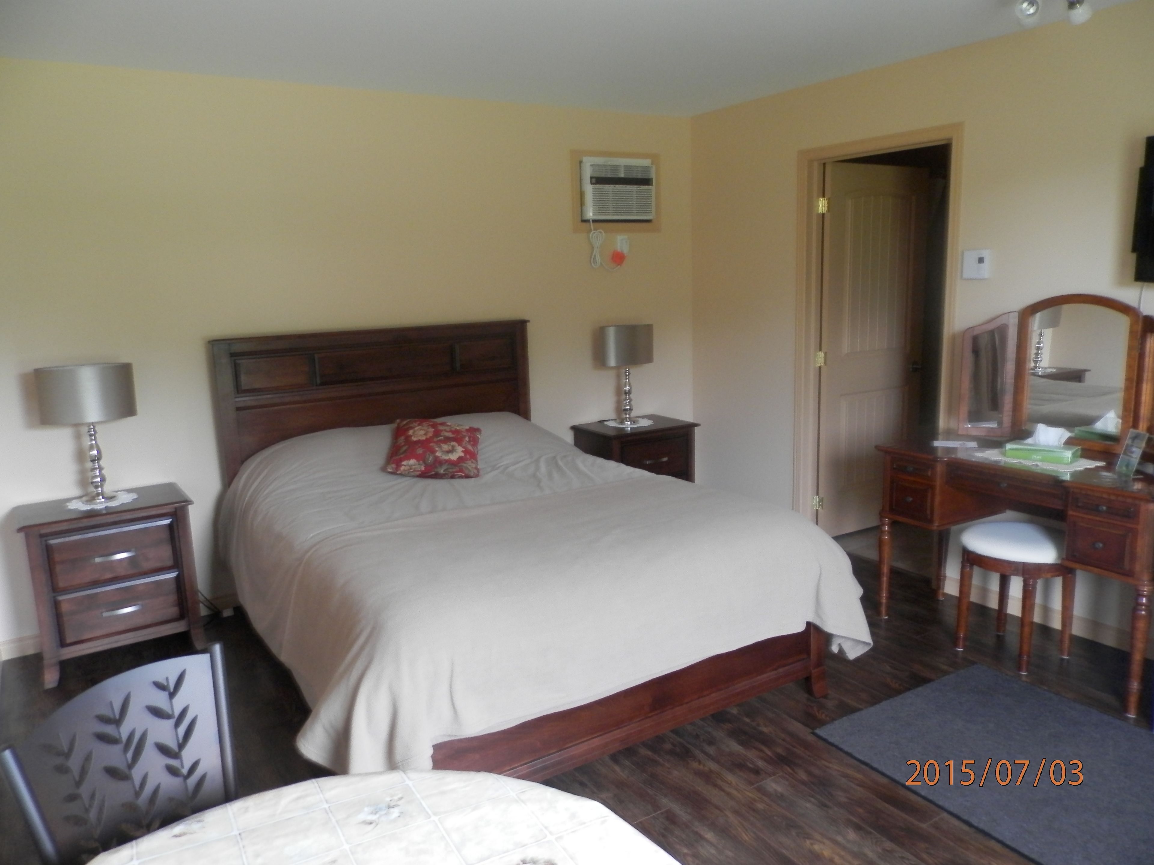 A bedroom with a bed, two bedside tables with lamps, a dresser with a mirror, and a door leading to another room.