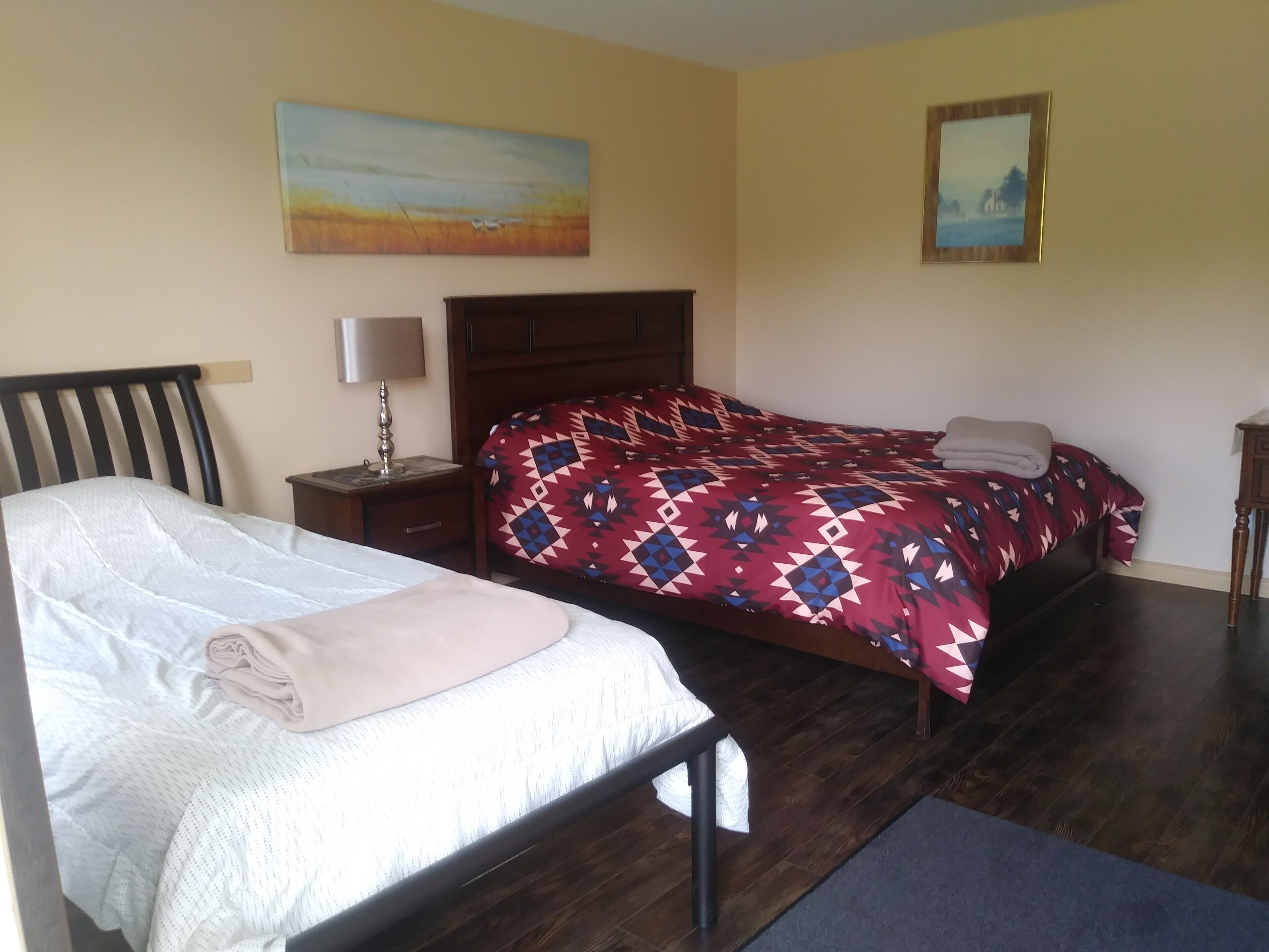 Een slaapkamer met een bed, nachtkastje, lamp, houten hoofdbord, een bed met een patroon en twee schilderijen aan de muur.