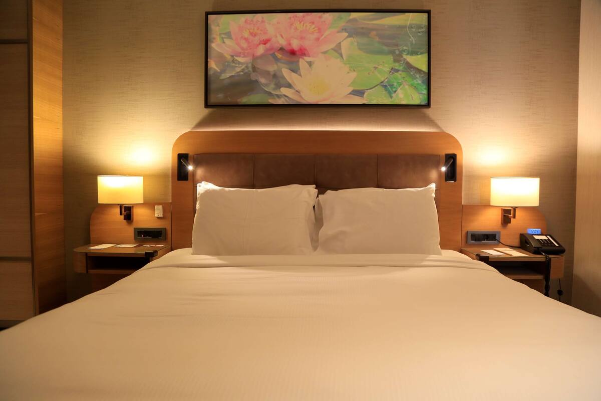 A hotel room with a large bed, two bedside tables with lamps, a wall-mounted painting of flowers, and a wooden headboard.