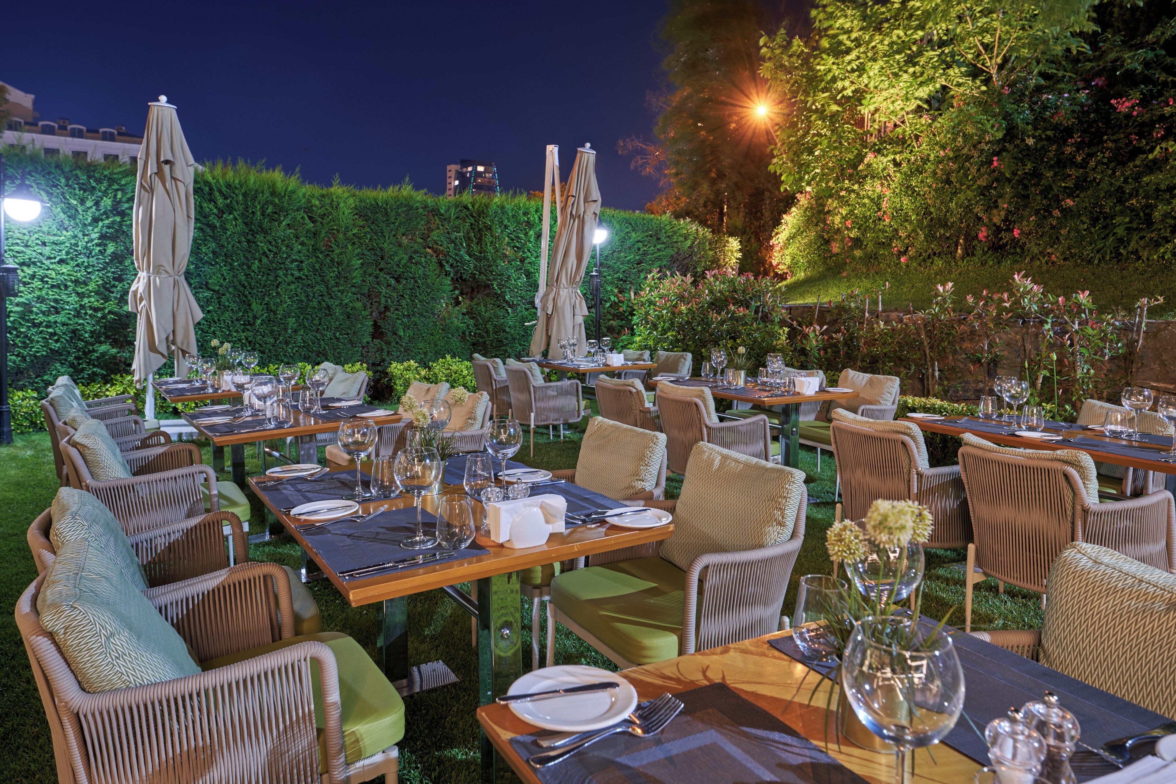 Un área de comedor al aire libre con mesas y sillas, rodeada de follaje y setos.