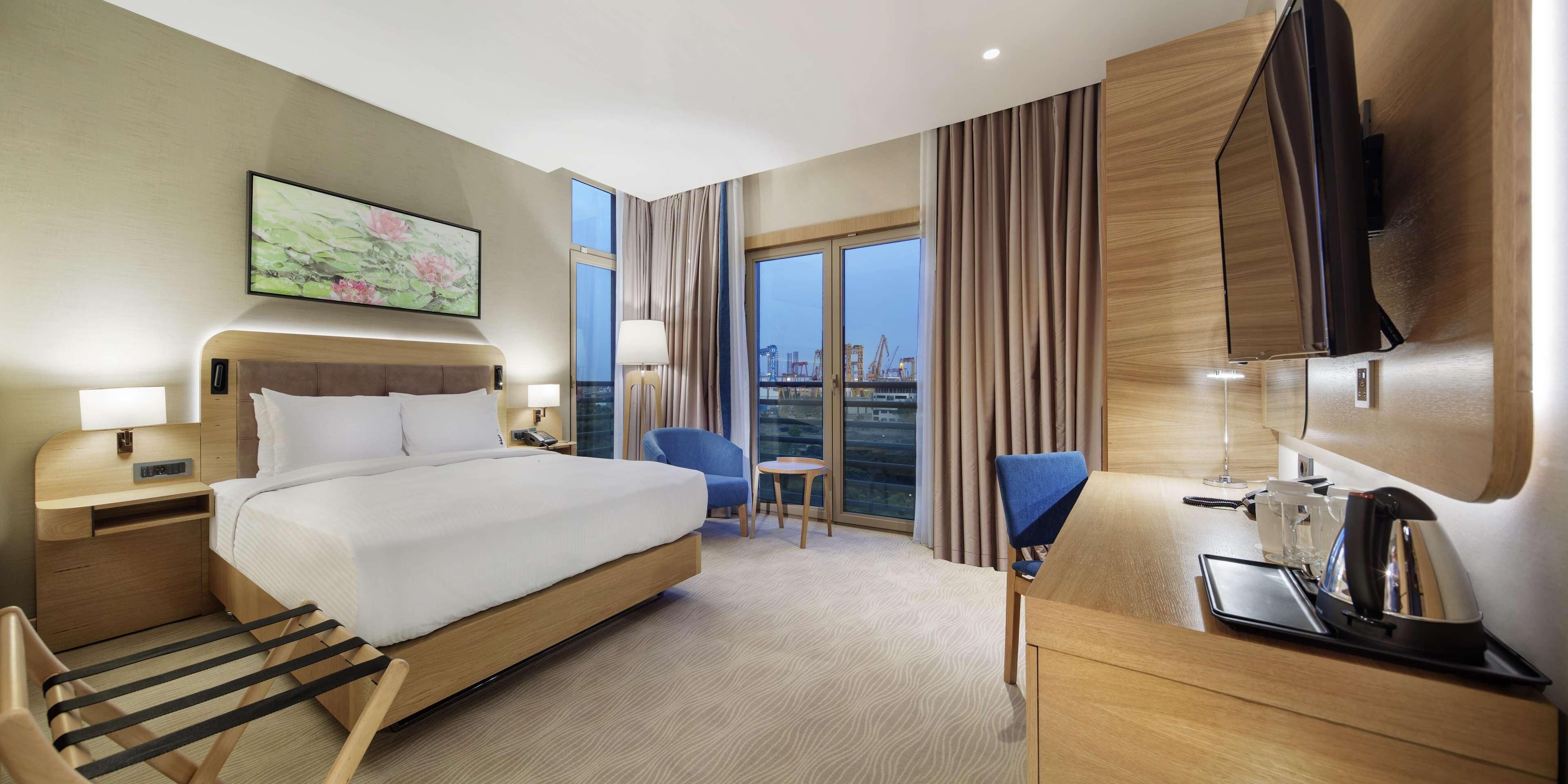 A hotel room with a bed, bedside tables, a desk with a kettle, a chair, and a large window with curtains.