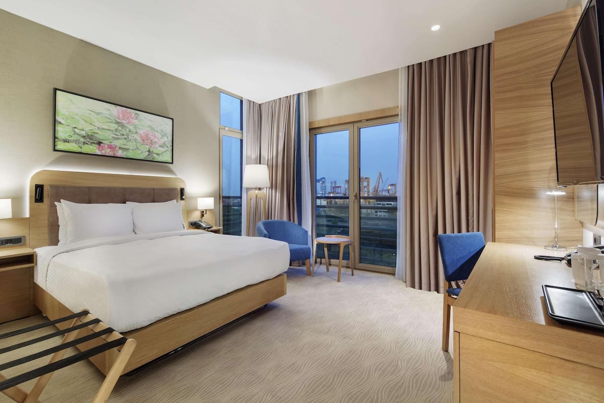 A hotel room with a bed, bedside tables, a desk with a kettle, a chair, and a large window with curtains.
