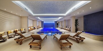 Un área de piscina interior moderna con sillones reclinables, una piscina central y un techo con tragaluces.