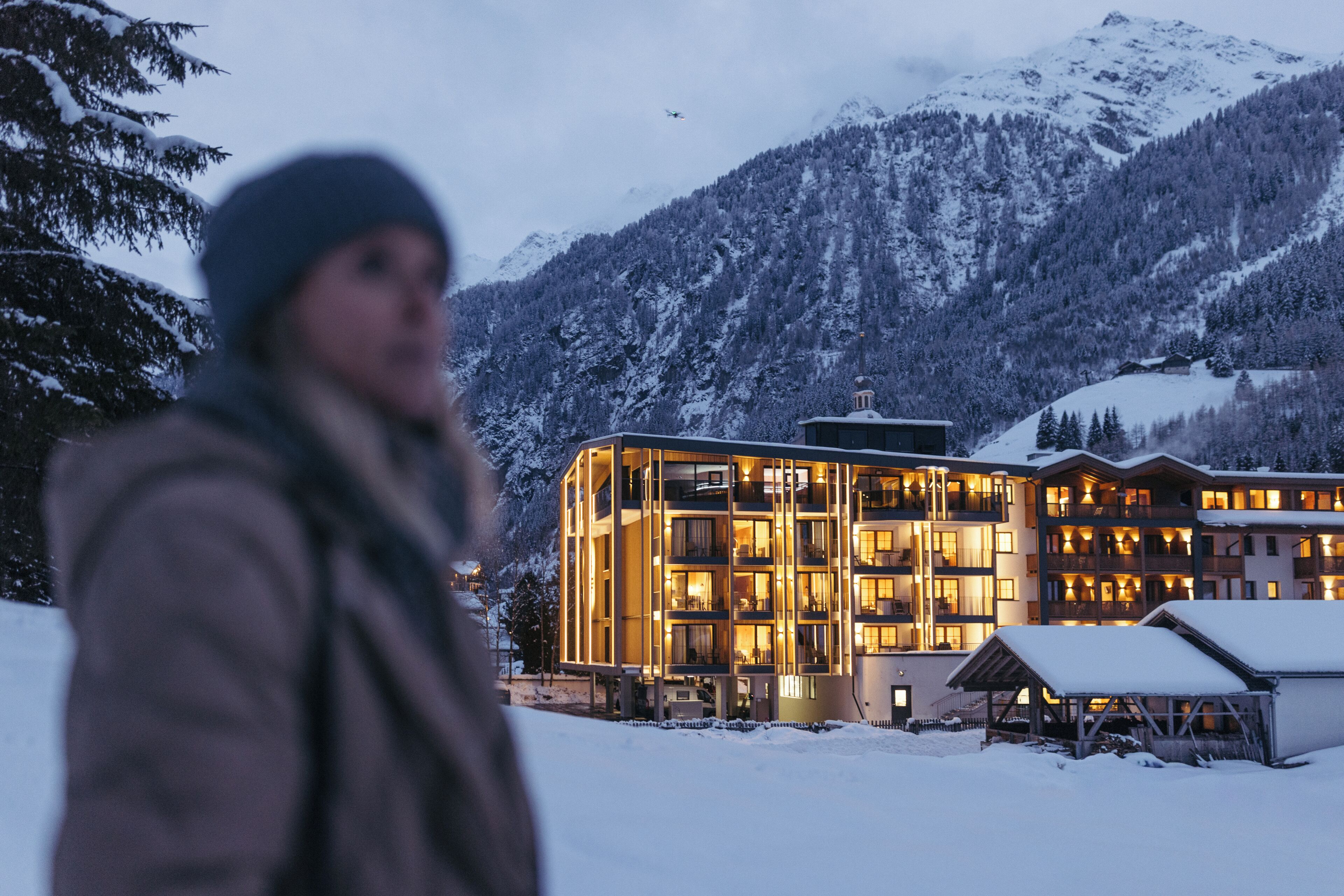 Un hotel de varios pisos con diseño moderno, con balcones y amplias ventanas, con una imponente estampa de montañas nevadas al fondo.