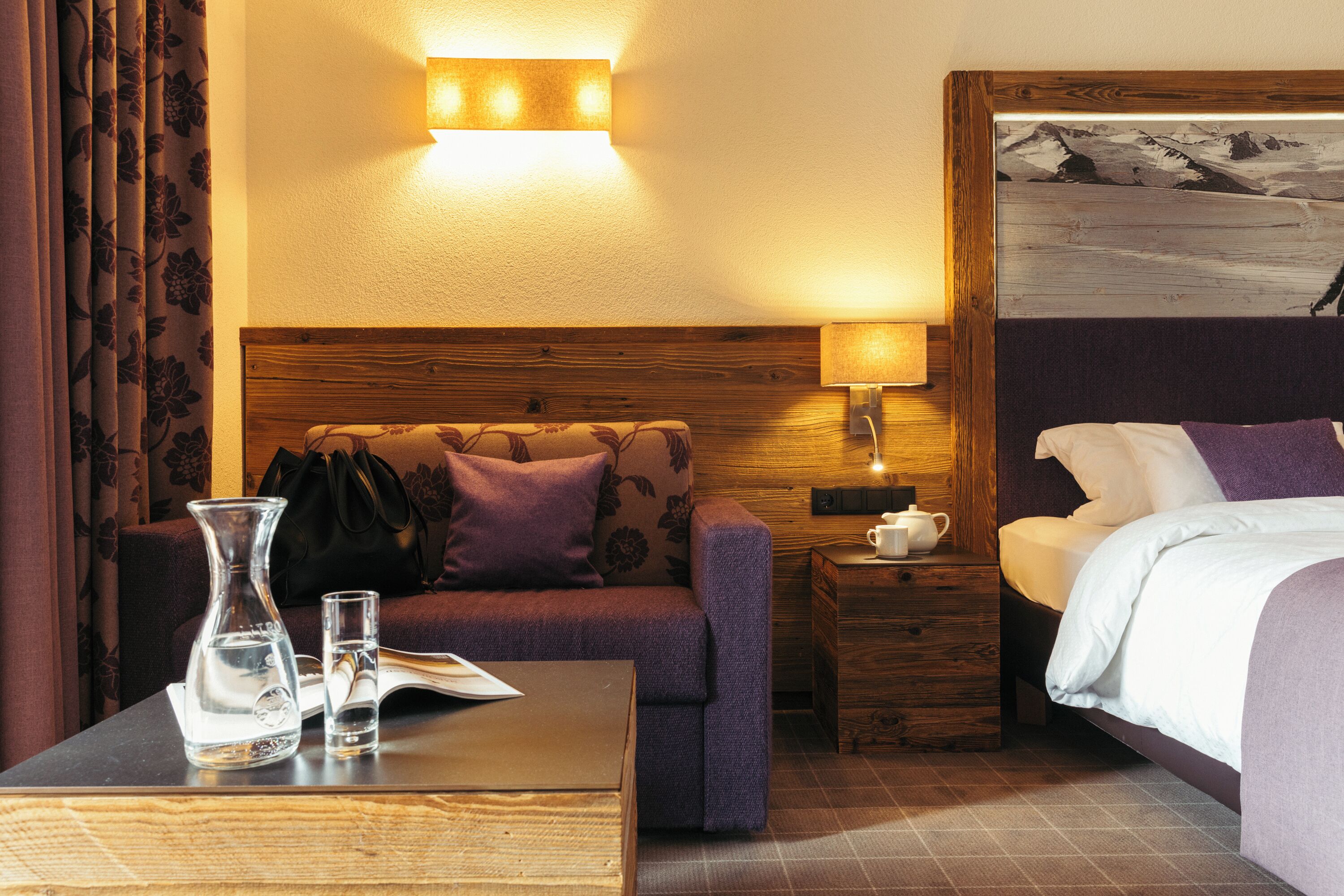 A hotel room with a wooden headboard, a sofa, a bed, a nightstand, a lamp, a painting, a window with curtains, and a table with a vase and glasses.