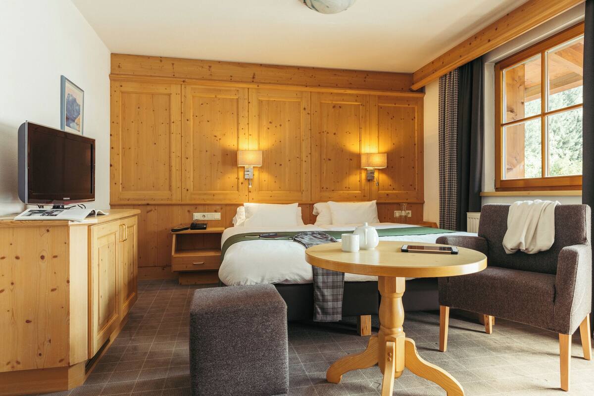 A hotel room with wooden paneling, a bed with white linens, a bedside table with a lamp, a television, and a window with curtains.