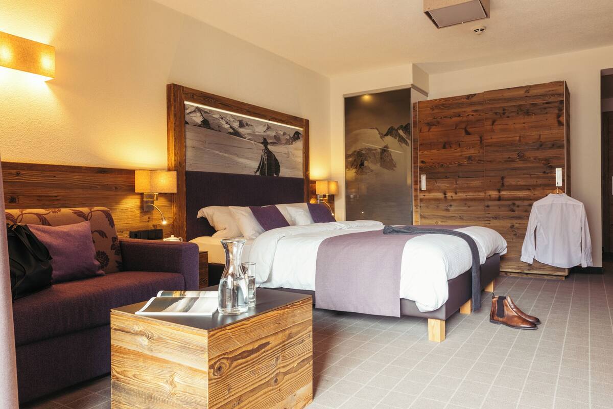 A hotel room with a large bed, a sofa, a wooden side table, and a wall-mounted TV.