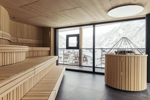 Un interior moderno con una zona de estar con banco de madera, un ventanal con vistas a montañas nevadas y una estructura redonda de madera con armazón metálico.