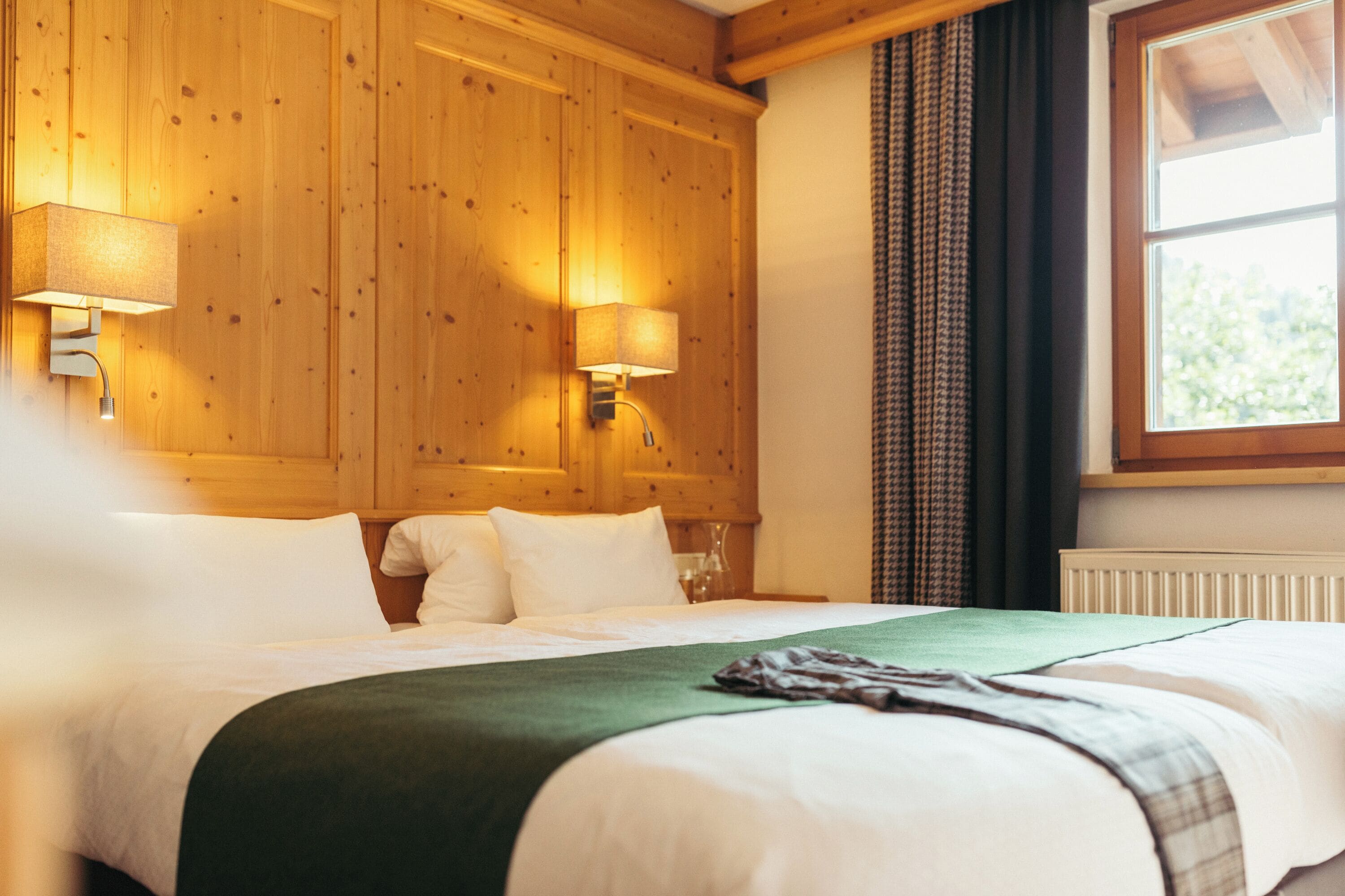 Habitación de hotel con cabecera de madera, dos lámparas de noche, un ventanal con cortinas y una cama bien tendida con sábanas blancas y una manta verde.