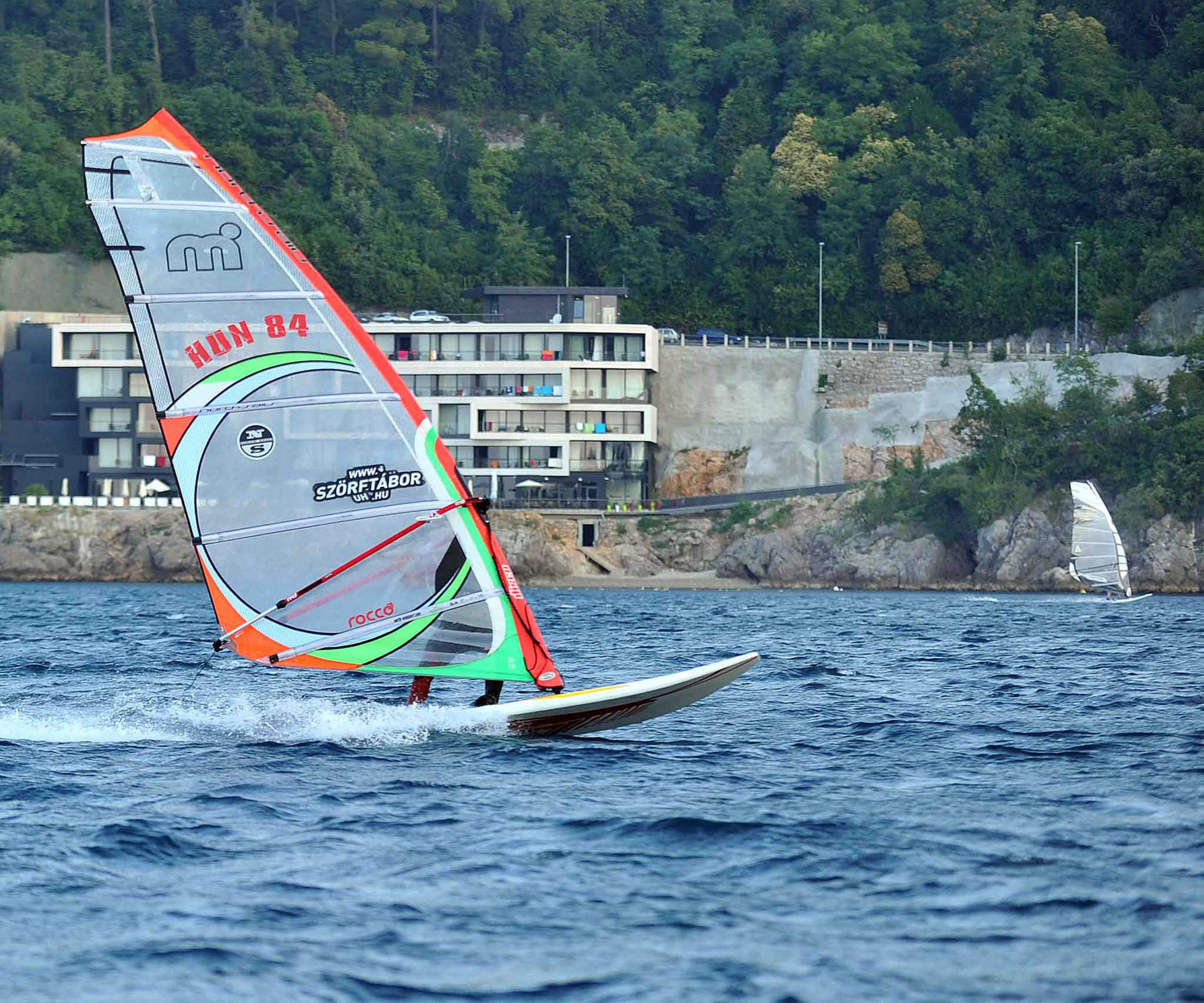 Un voilier aux voiles colorées sur l’eau, un bâtiment doté de balcons en arrière-plan et une côte rocheuse.