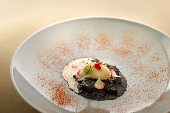 Un postre en un plato con borde blanco, decorado con una hoja verde y una flor roja, y espolvoreado con un polvo rojizo.