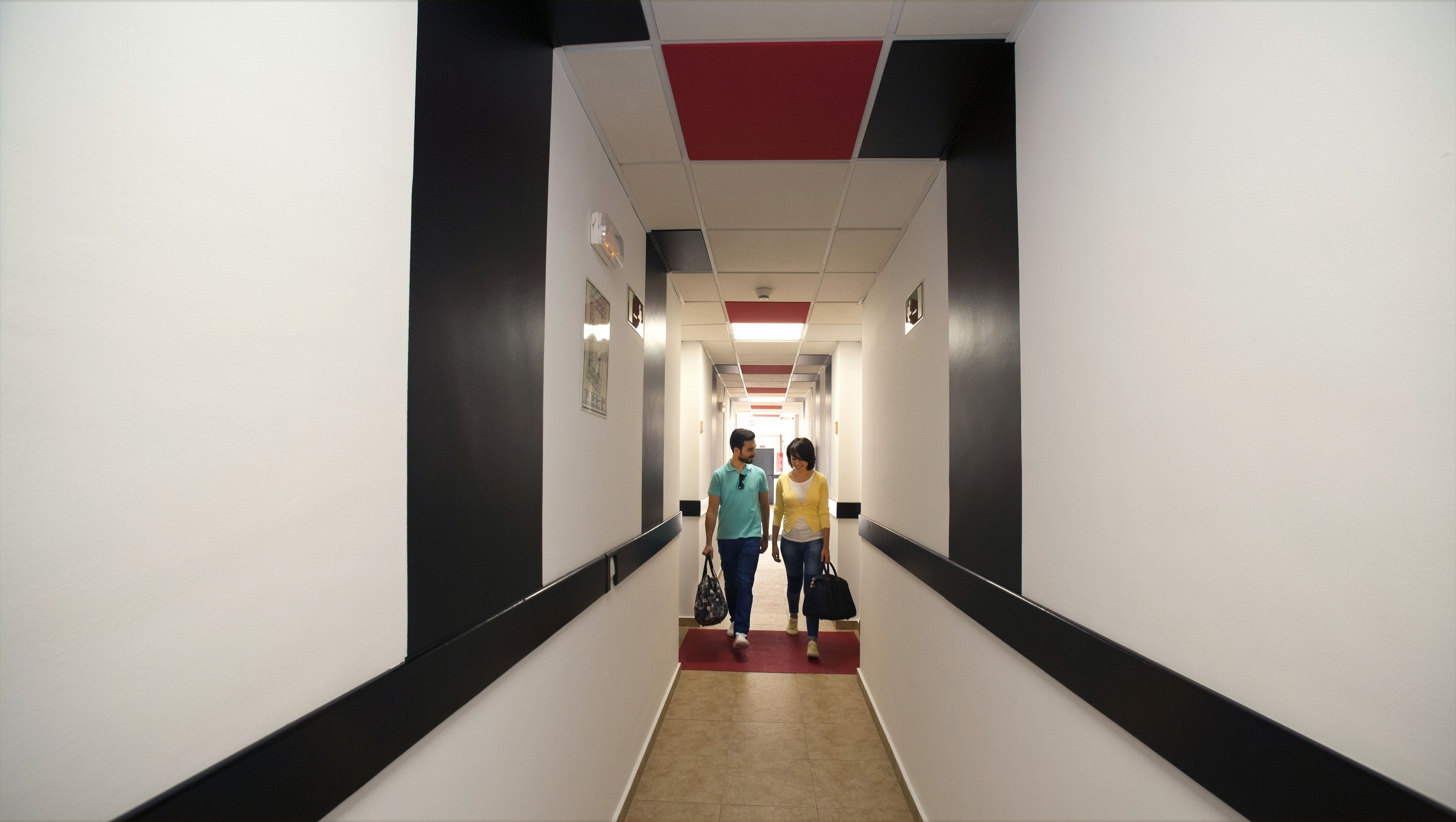 A hotel corridor with a red carpet, black and white walls, and a ceiling with red and white panels.