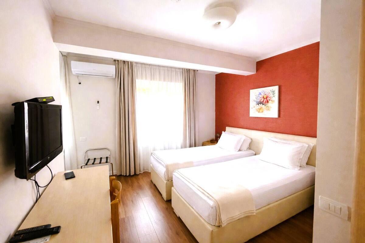 A hotel room with two beds, a TV, a desk, a chair, and a window with curtains.