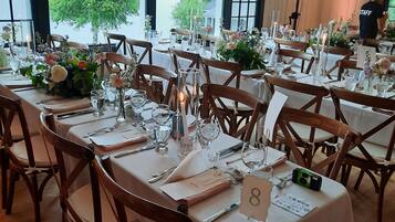 A restaurant with tables set for dining, large windows with a view of greenery, and a staff member in the background.