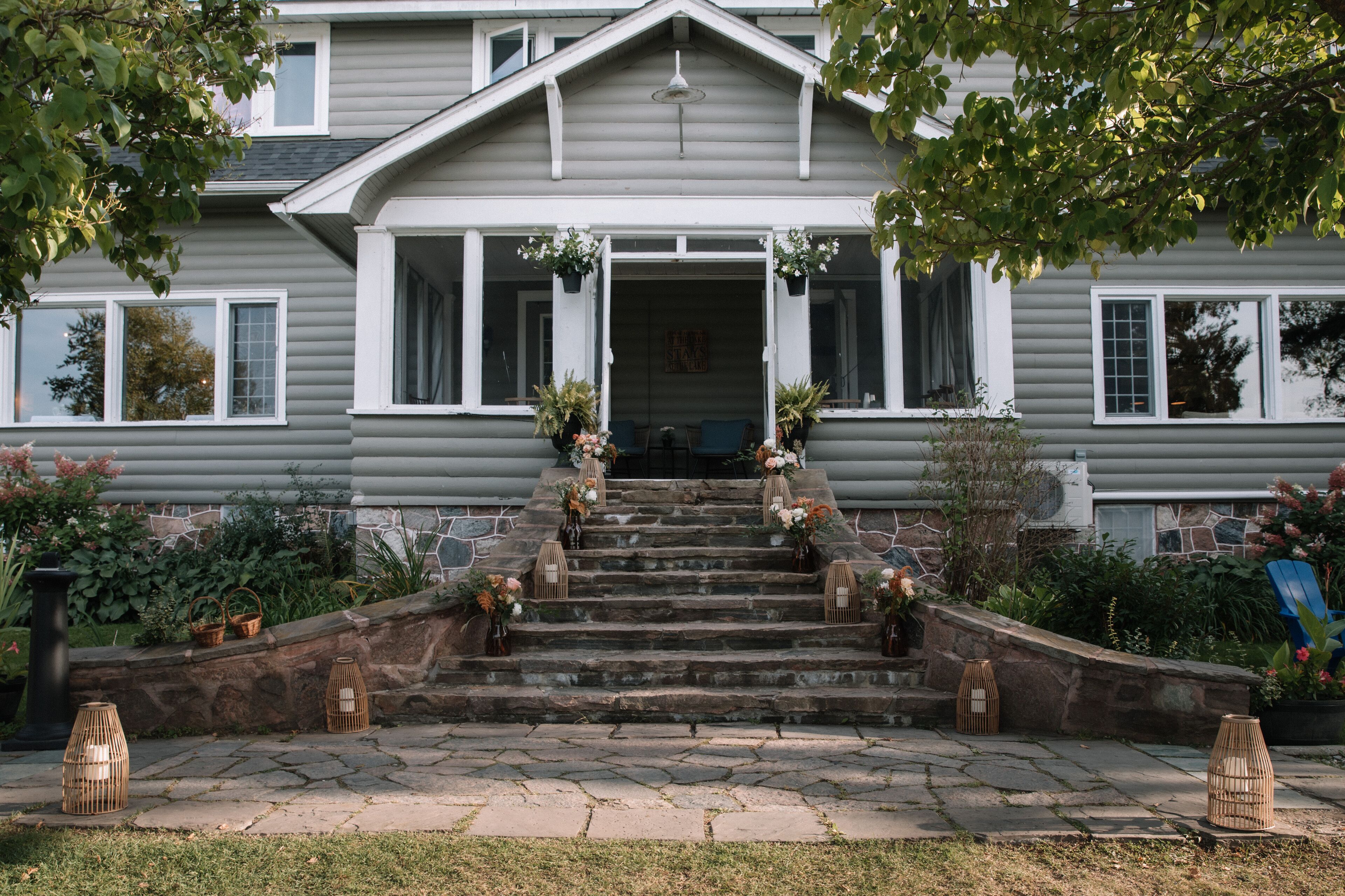 A two-story house with a stone staircase leading to the front door, surrounded by potted plants and outdoor furniture.