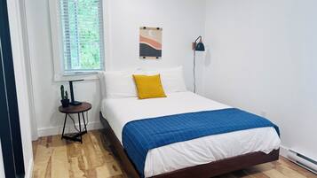 A neatly arranged bedroom with a bed, a small table with a lamp, a window with blinds, and a wall-mounted light fixture.