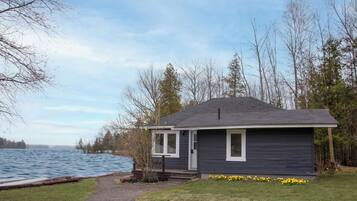 A small, dark-colored cabin by a lake with a dock, surrounded by trees.