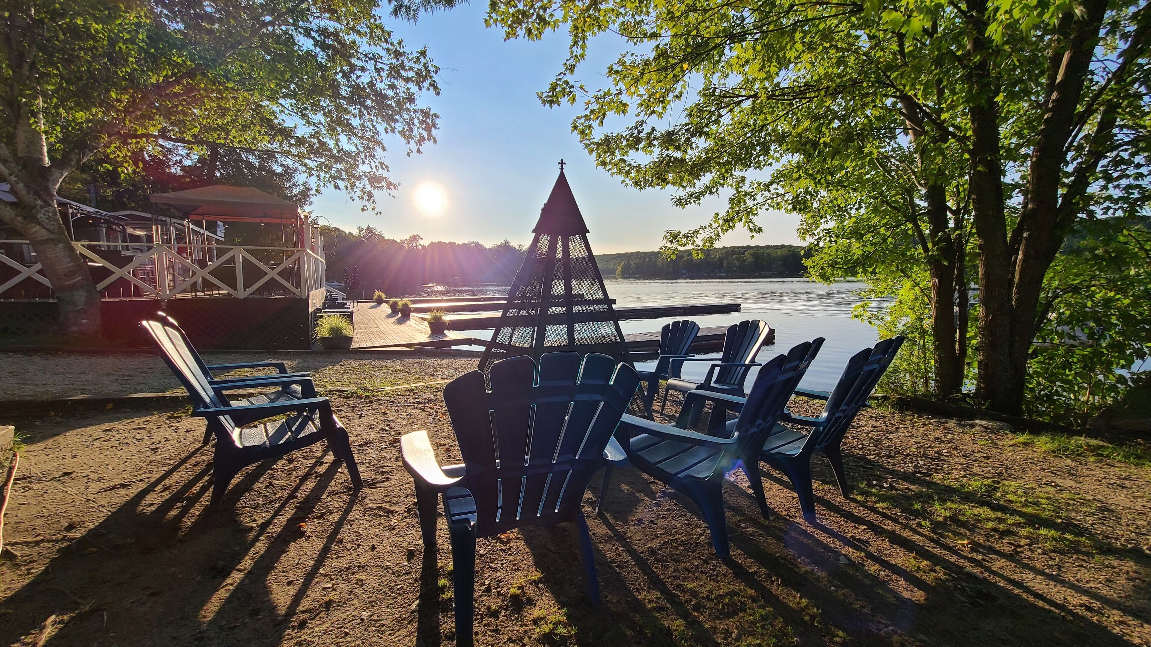 A lakeside seating area with Adirondack chairs and a table, surrounded by trees and a view of the lake.