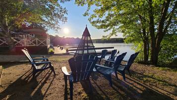 A lakeside seating area with Adirondack chairs and a table, surrounded by trees and a view of the lake.