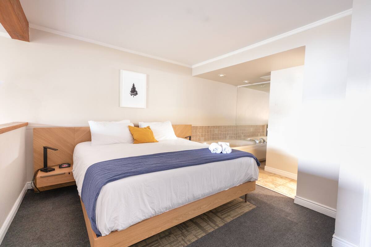 A modern hotel room with a large bed, a bedside table with a lamp, a small desk, and a bathroom visible through an open door.