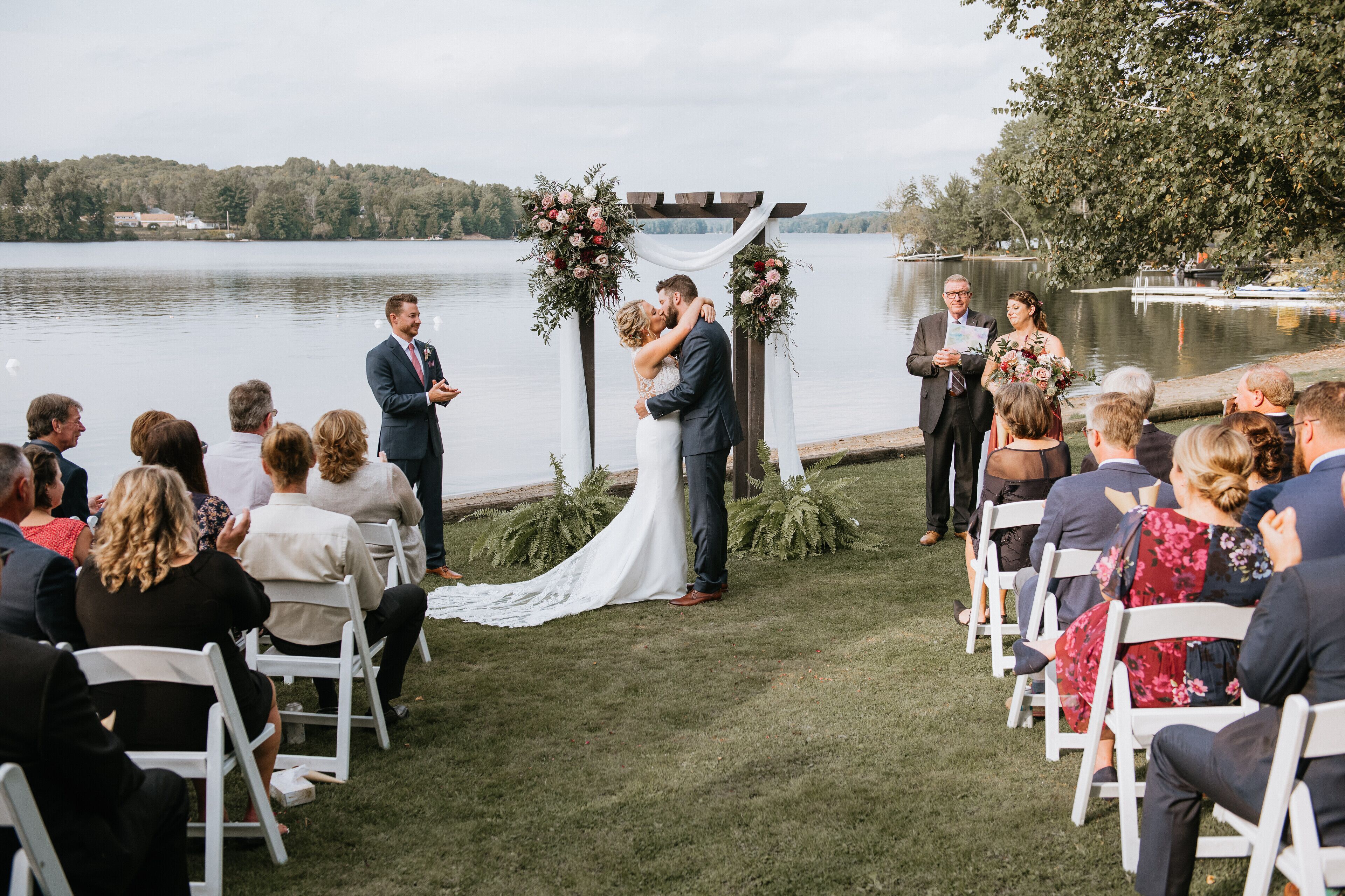 A wedding ceremony by a lake with guests seated on white chairs.