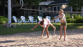 Two people playing volleyball on a sandy court with white chairs and a house in the background.