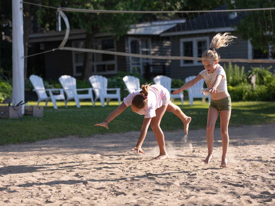 Two people playing volleyball on a sandy court with white chairs and a house in the background.
